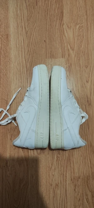 Nike Air Force 1 vita sneakers - Nike Air Force 1 sneakers i helvitt med klassisk låg siluett och rund tå. Skorna har snörning, perforerad tåbox och tydlig Nike Swoosh på sidorna. Tillverkade i skinn med platt sula och metall-detalj vid snörningen. Perfekt för dig som gillar clean och tidlös streetstil.