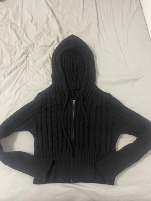 Svart stickad hoodie med dragkedja , STL L - Svart stickad hoodie med grova kabelstickade detaljer och ribbad nederkant. Tröjan har huva med snörning och dragkedja framtill. Långärmad och i en lite kortare modell som ger en chill vibe. Perfekt för dig som gillar mysiga och stilrena plagg.