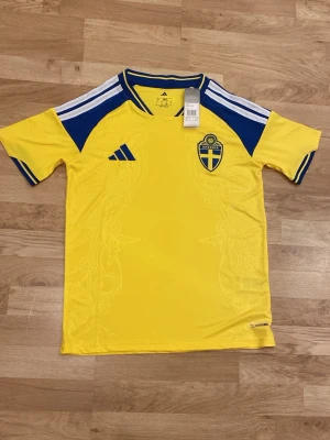 Sverige hemmatröja 25/26 - Sveriges nya hemmatröja i Storlek S. Tröjan är helt ny och har adidas samt landslagsmärke på bröstet. Vita ränder på axlarna och material som andas. Perfekt till VM-kvalet. 