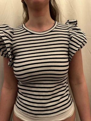 Randig topp med volangärmar - Säljer en vit och svart randig topp med korta volangärmar. Toppen har rund halsringning och är ribbad i materialet vilket ger en snygg struktur. Passformen är normal och toppen är perfekt för dig som gillar en lekfull och trendig stil.