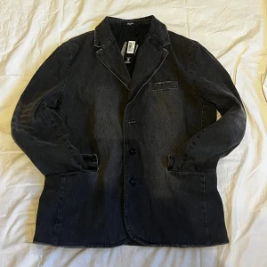 Jacka från Jaded London - Säljer en svart jeanskavaj från Jaded London med klassisk krage, två knappar framtill och tre fickor. Jackan har en overzised passform och är tillverkad i robust denim. Perfekt för dig som gillar en cool och stilren look. Jackan är aldrig använd