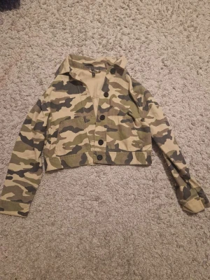 Camouflage croppad jacka H&M Divided S - Snygg croppad jacka från H&M Divided i camouflage-mönster. Jackan har knappar framtill, två stora fickor och är i bomullstyg. Perfekt för dig som vill sticka ut med en streetstyle-look.