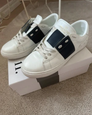 Valentino Garavani vita sneakers - Snygga vita sneakers från Valentino Garavani med bred marinblå detalj över sidan och vita nitar baktill. Skorna har snörning, rund tå och är tillverkade i skinn. Perfekta för dig som vill ha en clean men lyxig look.