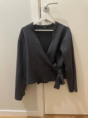 Mörkgrå omlottkofta från Gina Tricot - Snygg mörkgrå omlottkofta från Gina Tricot i mjukt stickat material. Koftan har v-ringad hals, långa ärmar och knyt i sidan för en cool och avslappnad look. Perfekt att slänga på sig när det är lite kyligt ute. På ena ärmen har den lite blekning på men de är så lite så de märks knappt. (jobbar som frisör)🥰🫶
