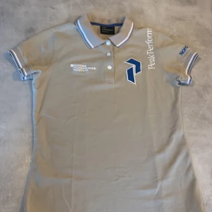 Beige Peak Performance polotopp M - Säljer en beige polotopp från Peak Performance i storlek M. Toppen har korta ärmar med blå och vita ränder på mudden, klassisk krage med tre knappar och broderade loggor i blått och vitt framtill. Materialet är bomull och toppen har en normal passform.