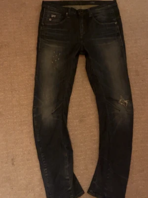 G-Star Raw denim jeans blå 27/32 - Snygga mörkblå jeans från G-Star Raw med klassisk femficksdesign och slitna detaljer på benen. Modellen har raka ben och normal passform, med coola sömmar och logopatch fram. Perfekt för dig som gillar streetstyle och vill ha ett par jeans med attityd.