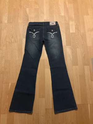 Mörkblå vintage jeans - Snygga vintage mörkblå jeans med låg midja och flare-ben. Baksidan har coola fickor med silverfärgade paljettdetaljer och knappar. Jeansen är tillverkade i klassiskt denim och har slitningar framtill för en trendig look.