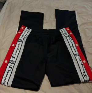 Champion svarta träningsbyxor XS - Säljer ett par svarta träningsbyxor från Champion i storlek XS. Byxorna har vita och röda sidopaneler med tryckta loggor och text, samt knappar längs benen för att kunna öppna upp. Elastisk midja och sportig look, perfekta för chill eller gym.Finns engast lite smutsigt längst nere på sista bild vissade.