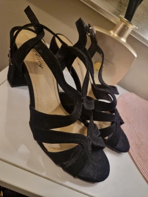 Svarta sandaler med klack från Alley - Svarta sandaler från Alley med bred klack och snygga korsade remmar i mockaimitation. Skorna har öppen tå och justerbart spänne vid ankeln. Perfekta för dig som vill ha en stilren och trendig look med lite höjd.