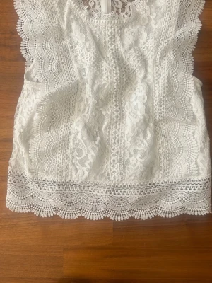 Vit spetsblus med volangdetaljer - Supersöt vit blus i spets med vackra volangdetaljer längs ärmhålen och nederkanten. Blusen har ett genomarbetat spetsmönster och rund halsringning. Perfekt för dig som gillar romantisk och feminin stil.