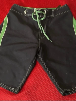 Ripzone svarta boardshorts med gröna detaljer - Ripzone boardshorts i svart med gröna och vita detaljer längs sidorna och bak. De har snörning i midjan och en bakficka med knapp. Materialet är 100% polyester som torkar snabbt, perfekt för strand och vattenaktiviteter. Snygga kontrastsömmar och sportig look.