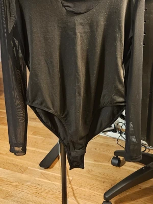Svart mesh body med lång ärm - Säljer en svart body med långärmade meshärmar och rund hals. Materialet är tunt och stretchigt, vilket ger en snygg och smickrande passform. Perfekt att styla med höga jeans eller kjol för en trendig look.