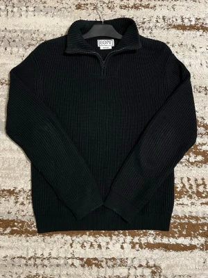 Svart stickad half zip-tröja CORE - Svart stickad tröja från CORE med ribbad struktur och half zip-dragkedja vid kragen. Modellen har långärmade ärmar och är tillverkad av återvunnet material. Perfekt för dig som gillar stilrena och enkla plagg med lite edge.