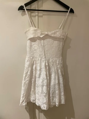 Vit spetsklänning med smala axelband - Söt vit kortklänning med smala axelband och blommig spets över hela klänningen. Klänningen har en lätt utställd kjol och en volangdetalj vid bysten. Perfekt för sommarens alla tillfällen!