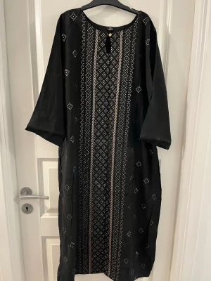 Svart långklänning med broderi XL - Svart långklänning i storlek XL med vackert broderade mönster i silver och beige längs hela framsidan. Klänningen har lång ärm, rund halsringning och en dekorativ knapp framtill. Materialet känns lätt och har en elegant glans.