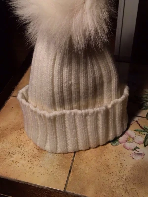 Vit stickad mössa med pälsboll - Säljer en vit ribbstickad mössa med stor fluffig pälsboll på toppen. Mössan har bred uppvikt kant och är gjord i mjukt stickat material som håller dig varm. Perfekt för kalla dagar och ger en snygg touch till din vinterstil.