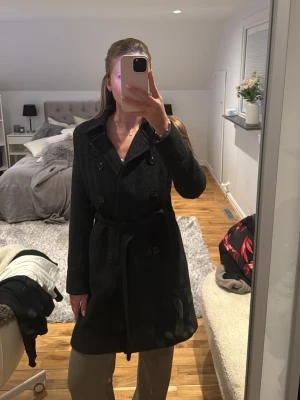 Svart kappa med bälte och knappar - Säljer en svart kappa med klassisk krage, dubbelknäppning och bälte i midjan. Jackan har en stilren och tidlös design som passar perfekt till kyligare dagar. Långärmad modell med snygg passform och diskreta detaljer.