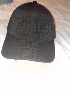 Svart Fendi keps  - Säljer en svart keps från Fendi med det klassiska FF-mönstret ton-i-ton över hela kepsen. Justerbar rem bak med Fendi-logga och snygga detaljer invändigt. Perfekt accessoar för dig som gillar exklusiv streetwear.