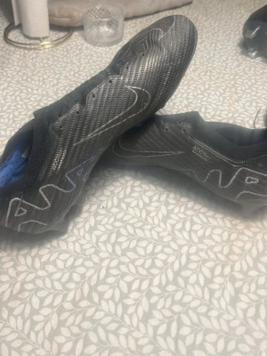 Nike Mercurial Vapor fotbollsskor svart - Säljer ett par svarta Nike Mercurial Vapor fotbollsskor med blå detaljer och Zoom-sula. Skorna har en lätt och syntetisk ovandel med ACC-teknologi för bästa bollkontroll. Mönstrad yta och klassisk snörning. Perfekta för dig som vill ha speed på planen.