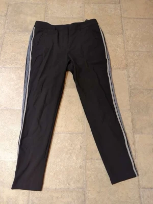 Svarta byxor med revärer från Vero Moda - Svarta byxor från Vero Moda i storlek M med grå revärer längs sidorna. Byxorna har en slim passform och är midwaist. Perfekta för en trendig och sportig look.