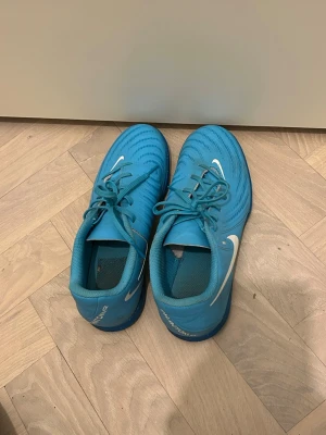 Nike Phantom blå fotbollsskor 38 inomhus - Säljer ett par Nike Phantom inomhus fotbollsskor i klarblå färg med vita detaljer och loggor. Skorna har snörning och en rund tå, samt en platt sula med mönstrad undersida för bra grepp. Materialet är syntet och skon har en sportig design som sticker ut på planen. Jättebra skick, storlek 38.
