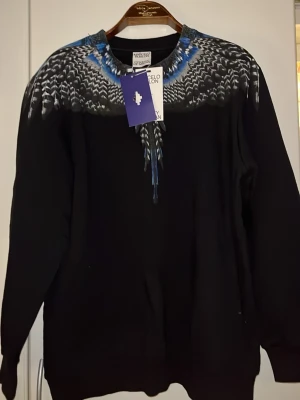 Svart tröja med blått fjädermönster Marcelo Burlon - Säljer en svart långärmad tröja från Marcelo Burlon med ett unikt blått och vitt fjädermönster runt halsen. Tröjan har en relaxed passform och är tillverkad i mjuk bomull. Perfekt för dig som vill sticka ut med cool design.