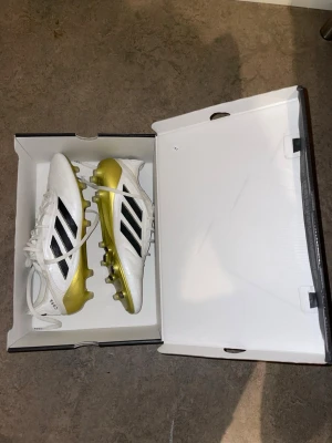 Adidas Copa fotbollsskor vit/guld - Säljer ett par Adidas Copa fotbollsskor i vitt med guldiga detaljer och svarta ränder. Skorna har snörning, platt sula med dobbar och rund tå. Ovandelen är i syntetmaterial och insidan har lila foder. Perfekt för dig som vill sticka ut på planen.