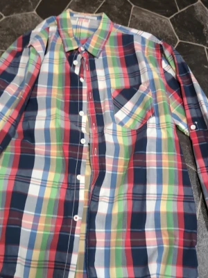 Flerfärgad rutig skjorta stl L - Säljer en färgglad rutig skjorta med knappar framtill och bröstficka. Skjortan har långa ärmar och klassisk krage. Perfekt för dig som gillar att sticka ut med din stil.