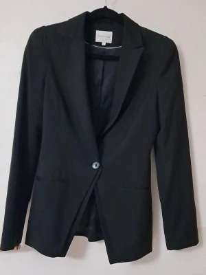 Svart kavaj från STOCKH LM - Klassisk svart kavaj från STOCKH LM med en knapp framtill och två fickor. Kavajen har slät yta, helfodrad insida med diskret logomönster och lång ärm. Perfekt för en stilren look och enkel att matcha med olika outfits.