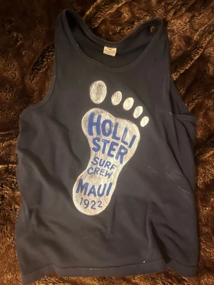 Mörkblå linnetopp från Hollister - Säljer en mörkblå linnetopp från Hollister i storlek S. Toppen har ett stort tryck med en fot och texten 'Hollister Surf Crew Maui 1922' i blått och vitt framtill. Perfekt för chill dagar eller strandhäng.
