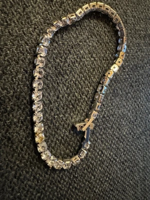 Glittrig silverfärgad tennisarmband - Säljer ett snyggt tennisarmband med glittriga stenar i silverfärg. Armbandet har en klassisk design med fyrkantiga länkar och gnistrande stenar som ger en lyxig känsla. Perfekt för att lyfta din outfit med lite extra bling.