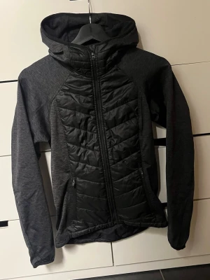 Svart sportig pufferjacka från H&M - Sportig svart pufferjacka från H&M med huva och hel dragkedja framtill. Jackan har quiltade paneler på bröstet och ryggen, samt två sidofickor med dragkedja. Materialet är en mix av polyester och syntet, perfekt för aktiva dagar.