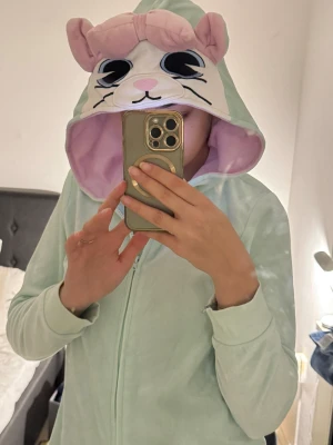 Mintgrön mjuk onesie med katt-hood - Supermysig mintgrön onesie med dragkedja och huva som har ett gulligt kattansikte, öron och rosett i ljusrosa. Insidan av huvan är ljuslila. Perfekt för chill hemma eller sleepover. Materialet är mjukt och känns som fleece.