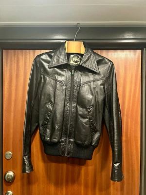 Croppad läderjacka 70-tal, bomber - En cool gammal läderjacka med croppad passform och stor krage a la 70-tal. Dragkedjan är lite wonky men funkar helt okej med rätta knycken. Bäst för någon 46-48