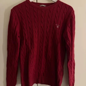 Vinröd kabelstickad tröja från GANT - Snygg vinröd kabelstickad tröja från GANT med rund hals och diskret logga broderad på bröstet. Tröjan har långa ärmar och ribbade muddar vid hals, ärmslut och nederkant. Perfekt för dig som gillar klassisk stil med en modern touch