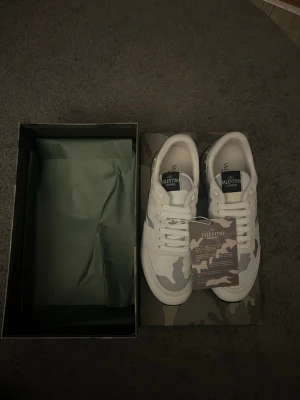 Valentino Garavani Rockrunner sneakers - Säljer ett par Valentino Garavani Rockrunner sneakers i vitt med grått och beige camouflage-mönster. Skorna har rund tå, snörning och platt sula. Materialet är en mix av skinn och textil. Kommer med originalkartong och dustbag. Perfekt för dig som gillar exklusiva sneakers med unik design.