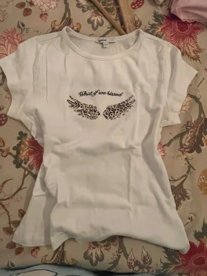 Vit t-shirt med leopardvingar Subdued - Vit t-shirt från Subdued i storlek M med tryck på bröstet: 'What if we kissed' och två leopardmönstrade vingar. T-shirten har rund hals och korta ärmar, tillverkad i mjuk bomull. Perfekt för dig som gillar coola prints och enkel stil.