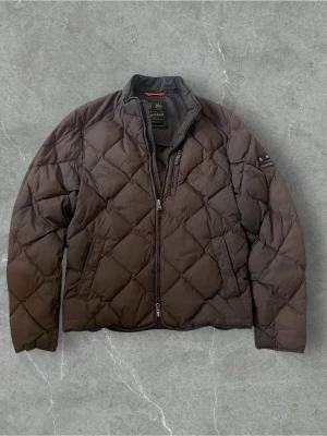Brun quiltad pufferjacka från Morris | S/M - Quiltad pufferjacka från Morris i mörkbrun färg med snyggt rutmönster. Jackan har hög krage, två sidofickor och grov dragkedja framtill. Materialet är polyester och jackan har en klassisk, normal passform. Perfekt för kyliga dagar och stilren look.