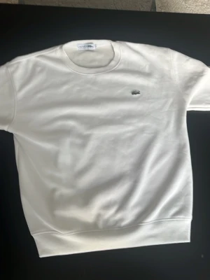 Vit sweatshirt från Lacoste - Säljer en stilren vit sweatshirt från Lacoste med klassisk rund hals och det ikoniska krokodilloggan broderad på bröstet. Tröjan har långa ärmar och ribbade muddar vid ärmslut och nederkant. Perfekt för en clean och enkel look.
