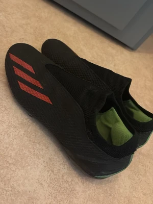 Svarta adidas fotbollsskor strl 41 - Säljer ett par svarta adidas fotbollsskor med röda detaljer och grön insida. Skorna har en strömlinjeformad design utan snörning och är tillverkade i syntetmaterial för bästa grepp på planen. Perfekta för dig som vill ha stilrena och funktionella skor till matchen.