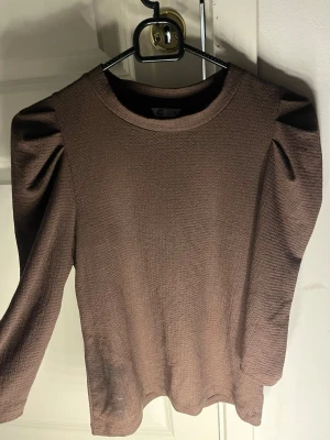 Brun långärmad blus med puffärm - Säljer en brun blus från C i storlek M. Toppen har lång ärm med snygga puffdetaljer vid axlarna och är tillverkad i ett strukturerat, mjukt tyg. Rund halsringning och loose passform ger en chill vibe. Perfekt att styla med jeans eller kjol.