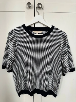 Topp i nyskick från Pernilla Wahlgren XS - Säljer en svartvit hundtandsmönstrad topp från Pernilla Wahlgren i storlek XS. Toppen har rund hals, korta ärmar och ribbade kanter i svart. Materialet är stickat och har en liten guldfärgad detalj längst ner. Som ny!