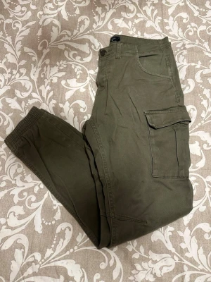 Olivgröna cargopants från Lager 157 - Säljer ett par olivgröna cargopants från Lager 157 i storlek S. Byxorna har elastiska muddar vid bensluten, stora fickor på sidorna och klassisk cargostil. Materialet är mjukt och känns som bomull, perfekt för en chill och avslappnad look.
