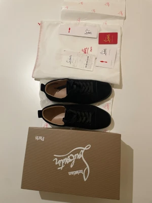 Svarta sneakers från Christian Louboutin  - Säljer ett par svarta sneakers från Christian Louboutin med klassisk röd sula och snygg snörning. Skorna är i mocka och har en rund tå. Medföljer originalkartong, dustbag och äkthetskort. Perfekt för dig som gillar exklusiva detaljer och stilren design. 100% äkta med kvitto används ett fåtal gånger kolla bilderna.