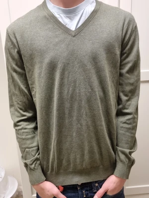 Olivgrön v-ringad tröja Riley L - Olivgrön v-ringad tröja från Riley i storlek L. Tillverkad i 100% bomull med mjuk och tunn känsla. Tröjan har långa ärmar och ribbade muddar vid ärmslut och nederkant. Perfekt för lager-på-lager och enkel att matcha med jeans eller chinos. Passar 186