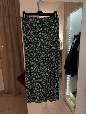 Svart grön blommig långkjol med slits - Säljer en långkjol med grönt och vitt blommönster. Kjolen har resår i midjan där bak och en slits på höger framsida. Från Lindex barn.