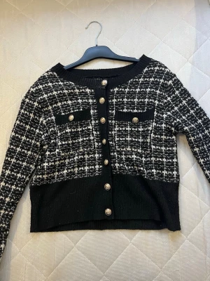 Svartvit rutig kofta med knappar - VERO MODA  - Snygg svartvit kofta med rutigt mönster och dekorativa guldfärgade knappar framtill. Koftan har två fejkade bröstfickor och breda svarta muddar vid nederkant och ärmslut. Perfekt för dig som gillar klassisk stil med en modern twist.