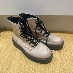 Beiga boots med blommigt mönster - Säljer ett par beiga boots med svart sula och snörning. Skorna har små blommiga detaljer i olika färger och en chunky platt sula. Perfekta för dig som vill ha något unikt och coolt till vardagslooken.