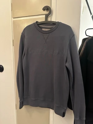 Mörkgrå sweatshirt från Calvin Klein Jeans - Snygg mörkgrå sweatshirt från Calvin Klein Jeans med broderad logga framtill. Tröjan har rund hals, ribbade muddar och långärmad passform. Perfekt för dig som gillar stilrena och klassiska plagg med en modern touch.