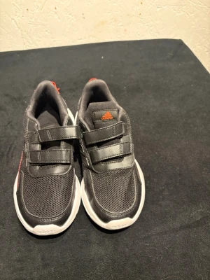 Svarta Adidas sneakers med kardborre - Säljer ett par svarta Adidas sneakers med vita sulor och klassiska tre ränder på sidan. Skorna har kardborreband för enkel på- och avtagning, mesh och syntetmaterial i ovandelen samt röda detaljer vid häl och logga. Perfekt för dig som gillar sportig stil.